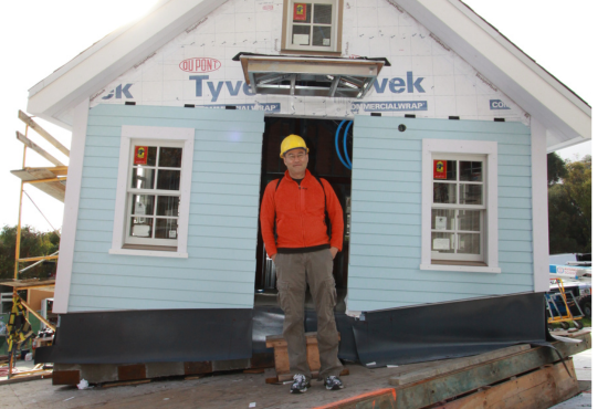 Do Ho Suh in front of Fallen Star while under construction, a blue house that was partially painted. Do Ho Suh is wearing a red sweater and dark pants and is smiling.  